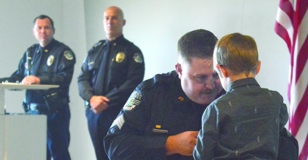 KYLE CRAVEN kneels as his son Matthew pins a sergeant’s badge on him. Capt. Robert Warner and Chief Greg Dull, in the background, lead the July 27 promotion ceremony, advancing four officers from the rank of corporal to sergeant.